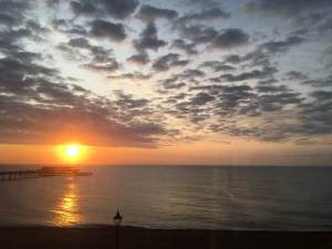 Sunset on a beach in England, with a calm sea and stratus clouds in the sky that is turning from blue to orange. A pier is in silhouette just beneath where the Sun is descending.